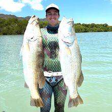 Load image into Gallery viewer, diver holding two large barramundi with water and trees in the background