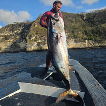 Load image into Gallery viewer, Man holding a large dogtooth tuna on a boat with a scenic background of mountains and water.
