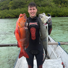 Load image into Gallery viewer, A man in a black wetsuit stands in a small boat holding two large fish, with green water and a forested shoreline in the background.
