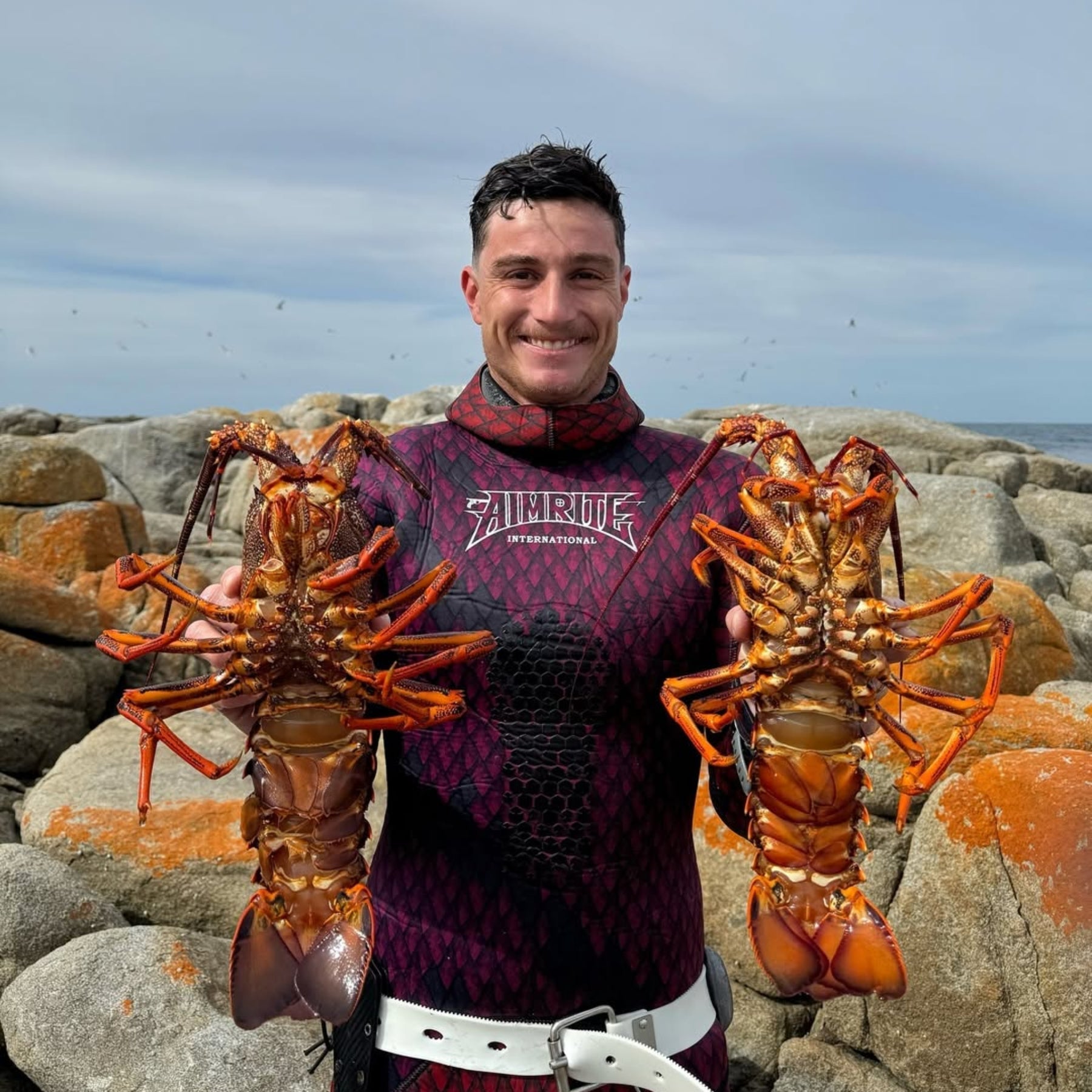 Aimrite spearo on the rocks holding two crayfish, wearing an Aimrite camo wetsuit